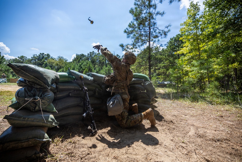 A soldier in full camouflage uniform and helmet is kneeling behind a sandbag fortification and throwing what appears to be a grenade or training device into the air. The scene takes place at an outdoor military training range surrounded by pine and deciduous trees. An M4/M16-style rifle is leaning against the sandbag wall beside the soldier.