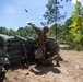 A soldier in full camouflage uniform and helmet is kneeling behind a sandbag fortification and throwing what appears to be a grenade or training device into the air. The scene takes place at an outdoor military training range surrounded by pine and deciduous trees. An M4/M16-style rifle is leaning against the sandbag wall beside the soldier.