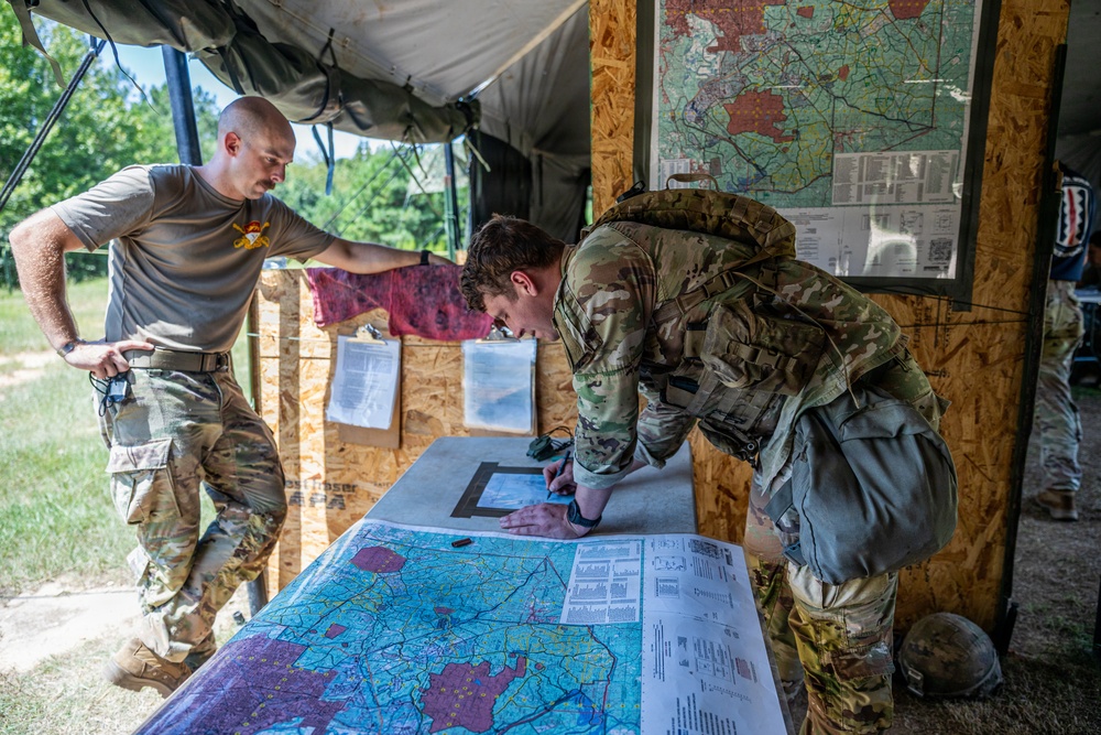 Two military personnel are working at an outdoor tactical operations center, with one soldier in full combat gear leaning over a large military map on a table while another in a t-shirt and combat pants observes. The setting appears to be a field command post with plywood walls, a canvas overhead cover, and additional maps posted on the wall. The scene suggests a planning or briefing activity during a military exercise or operation.