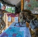 Two military personnel are working at an outdoor tactical operations center, with one soldier in full combat gear leaning over a large military map on a table while another in a t-shirt and combat pants observes. The setting appears to be a field command post with plywood walls, a canvas overhead cover, and additional maps posted on the wall. The scene suggests a planning or briefing activity during a military exercise or operation.