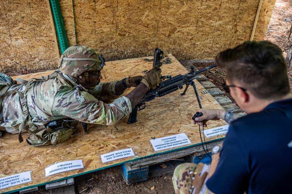 A soldier in full camouflage uniform and tactical gear is performing a weapons assembly or familiarization exercise on a plywood table surface. An evaluator or instructor in civilian clothing is observing and timing the exercise with a stopwatch. Labeled component cards are arranged on the table, indicating this is a formal weapons qualification or training event.
