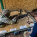 A soldier in full camouflage uniform and tactical gear is performing a weapons assembly or familiarization exercise on a plywood table surface. An evaluator or instructor in civilian clothing is observing and timing the exercise with a stopwatch. Labeled component cards are arranged on the table, indicating this is a formal weapons qualification or training event.