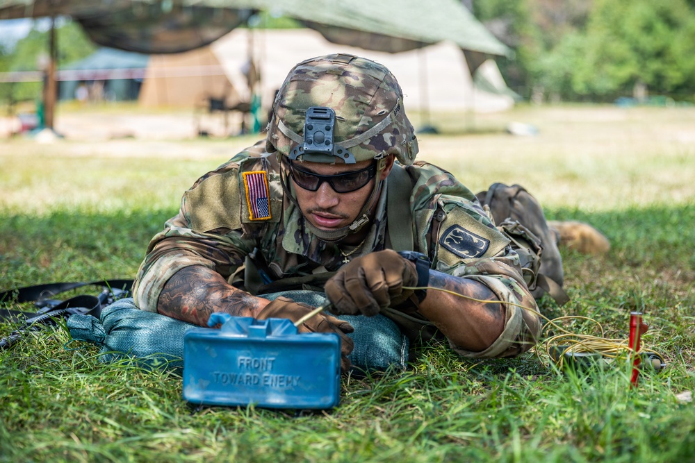 A U.S. Army soldier in full combat gear is lying prone on grass, working with a blue training Claymore mine marked 'FRONT TOWARD ENEMY.' The soldier is wearing OCP (Operational Camouflage Pattern) uniform with a U.S. flag patch and a unit patch, along with a combat helmet and tactical sunglasses. This appears to be a training exercise at a field camp, as indicated by the blue color of the device (denoting inert/training equipment) and the military tents visible in the background.