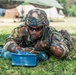 A U.S. Army soldier in full combat gear is lying prone on grass, working with a blue training Claymore mine marked 'FRONT TOWARD ENEMY.' The soldier is wearing OCP (Operational Camouflage Pattern) uniform with a U.S. flag patch and a unit patch, along with a combat helmet and tactical sunglasses. This appears to be a training exercise at a field camp, as indicated by the blue color of the device (denoting inert/training equipment) and the military tents visible in the background.