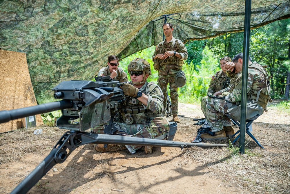 A group of military personnel in OCP (Operational Camouflage Pattern) uniforms are gathered under a camouflage net canopy in a wooded outdoor training area. One soldier in the foreground is positioned behind what appears to be an M2 .50 caliber heavy machine gun or similar crew-served weapon mounted on a tripod. The others appear to be observing or receiving instruction, suggesting a weapons familiarization or qualification training exercise.