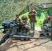 A group of military personnel in OCP (Operational Camouflage Pattern) uniforms are gathered under a camouflage net canopy in a wooded outdoor training area. One soldier in the foreground is positioned behind what appears to be an M2 .50 caliber heavy machine gun or similar crew-served weapon mounted on a tripod. The others appear to be observing or receiving instruction, suggesting a weapons familiarization or qualification training exercise.
