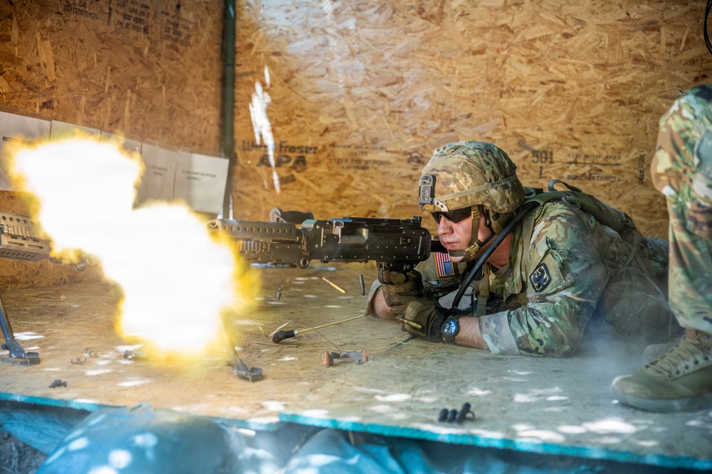 A U.S. Army soldier in full combat gear is firing a machine gun from a prone position inside a wooden shooting structure. A large muzzle flash is visible at the barrel, with spent brass casings ejecting to the right. This appears to be a live-fire training exercise at a military range.