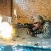 A U.S. Army soldier in full combat gear is firing a machine gun from a prone position inside a wooden shooting structure. A large muzzle flash is visible at the barrel, with spent brass casings ejecting to the right. This appears to be a live-fire training exercise at a military range.