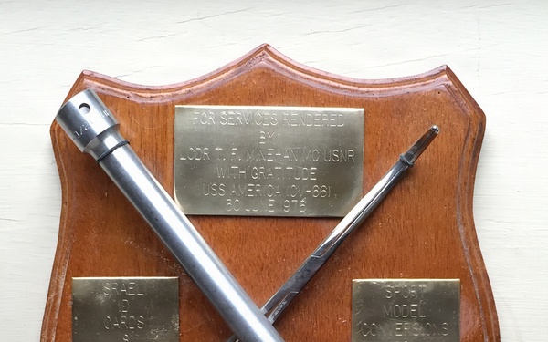 A wooden shield-shaped commemorative plaque presented to LCDR T.F. Minehan, MC USNR, for services rendered aboard USS America (CV-66) on 30 June 1976. Two medical instruments — a large syringe/injector and a hemostat/forceps — are mounted in a crossed pattern on the plaque. Three brass engraved plates are affixed to the wood, noting the dedication and what appear to be tally counts for procedures performed.