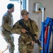 Two U.S. Air Force personnel in OCP (Operational Camouflage Pattern) uniforms are shown inside what appears to be a military facility or hangar. The foreground individual, wearing glasses and a patrol cap, is holding an M4-style carbine rifle with accessories. They appear to be conducting a training exercise or security patrol.