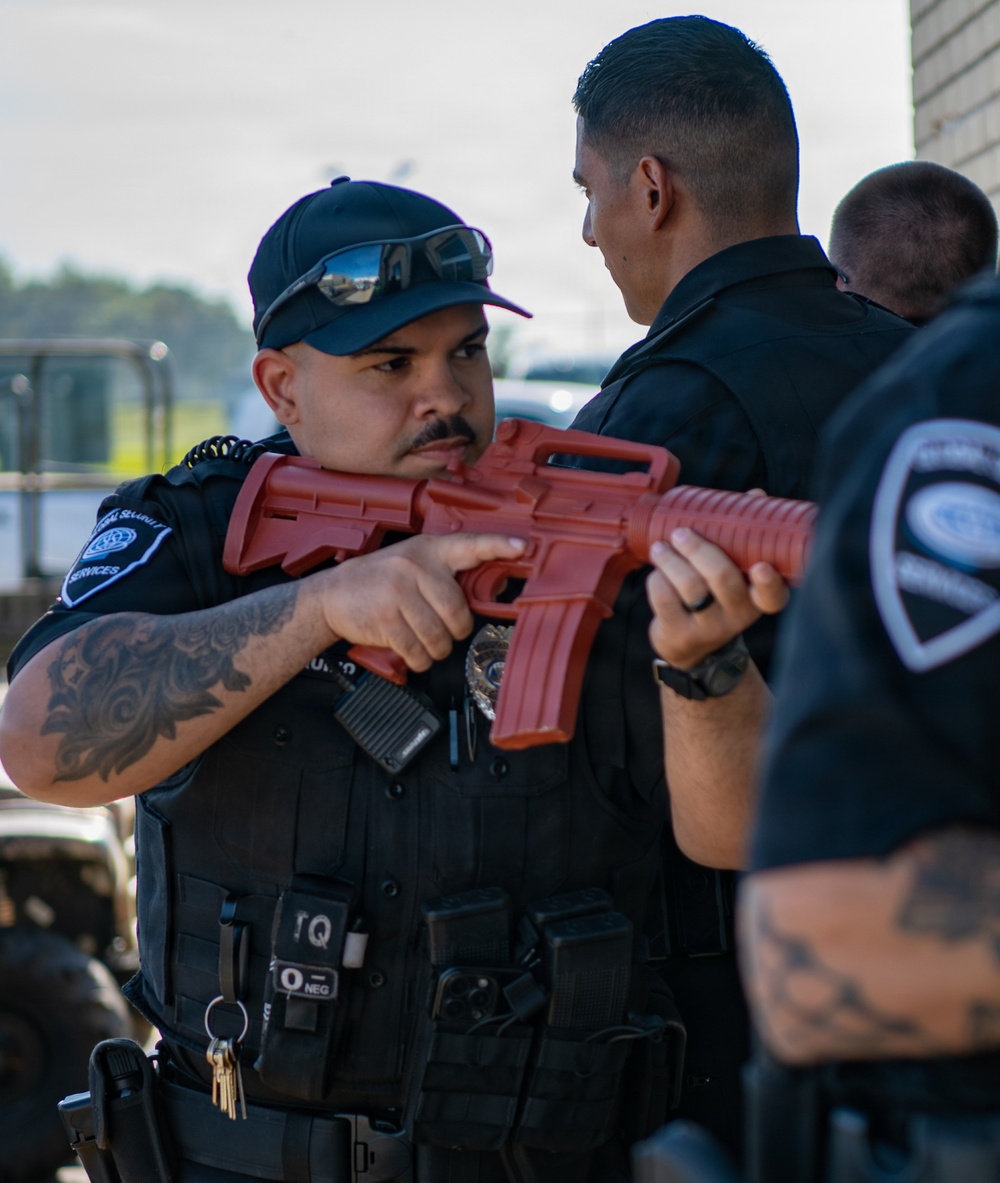 Two security personnel are engaged in what appears to be a training exercise, with one officer holding a red plastic/rubber training rifle replica. The officer in the foreground wears a tactical vest, baseball cap with sunglasses, and has visible sleeve tattoos. A third officer is partially visible in the right foreground, and all individuals wear matching dark uniforms with security service patches.