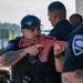 Two security personnel are engaged in what appears to be a training exercise, with one officer holding a red plastic/rubber training rifle replica. The officer in the foreground wears a tactical vest, baseball cap with sunglasses, and has visible sleeve tattoos. A third officer is partially visible in the right foreground, and all individuals wear matching dark uniforms with security service patches.