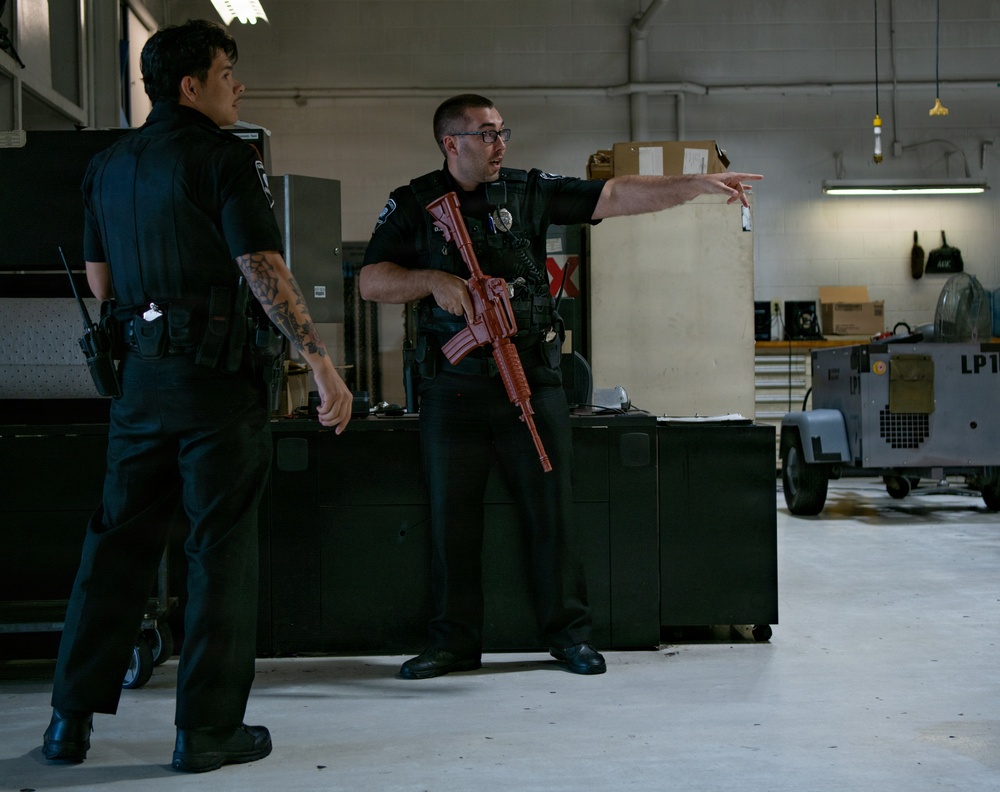 Two individuals in black law enforcement uniforms are inside what appears to be a police equipment bay or training facility. One officer is holding a red-colored training rifle (inert/dummy weapon used for training purposes) and pointing in a direction while appearing to give instructions. The setting includes storage equipment, shelving, and what appears to be a ground support vehicle in the background.