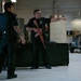 Two individuals in black law enforcement uniforms are inside what appears to be a police equipment bay or training facility. One officer is holding a red-colored training rifle (inert/dummy weapon used for training purposes) and pointing in a direction while appearing to give instructions. The setting includes storage equipment, shelving, and what appears to be a ground support vehicle in the background.