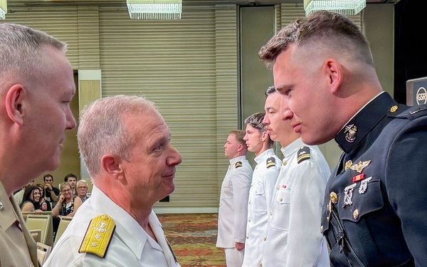 A military commissioning or awards ceremony is taking place in what appears to be a hotel ballroom or conference center. A senior naval officer in white uniform with gold shoulder boards is shaking hands with a Marine officer in dress blues, while a U.S. Marine general in tan service uniform observes from the left. Several naval officers in white uniforms stand at attention in the background.