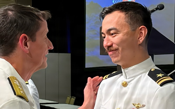 Two U.S. Navy officers in white dress uniforms are engaged in what appears to be a formal ceremony, likely a promotion or award presentation. The officer on the left is shaking hands with and pinning something onto the uniform of the officer on the right. The setting appears to be an indoor venue with a projection screen visible in the background.