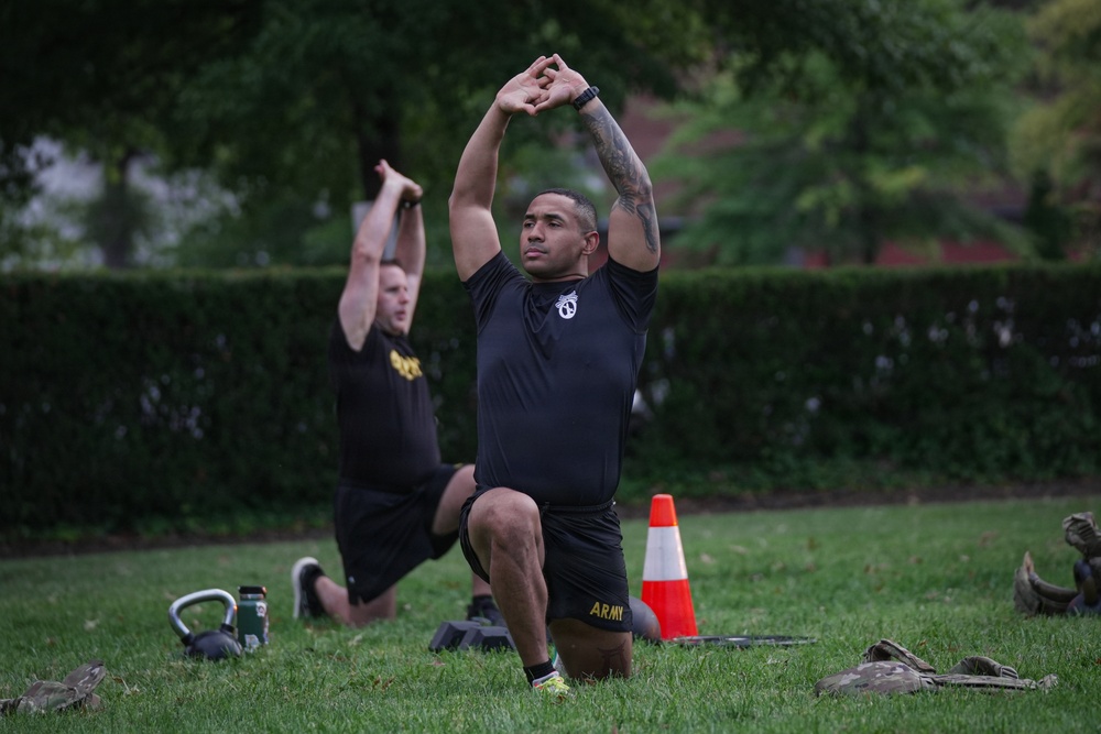 Two soldiers in U.S. Army physical training uniforms are performing lunge stretches with arms raised overhead on a grass field. The foreground soldier wears black shorts with 'ARMY' printed in gold lettering and has visible tattoos on his left arm. The setting appears to be an outdoor PT session with training equipment visible on the ground.