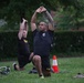 Two soldiers in U.S. Army physical training uniforms are performing lunge stretches with arms raised overhead on a grass field. The foreground soldier wears black shorts with 'ARMY' printed in gold lettering and has visible tattoos on his left arm. The setting appears to be an outdoor PT session with training equipment visible on the ground.