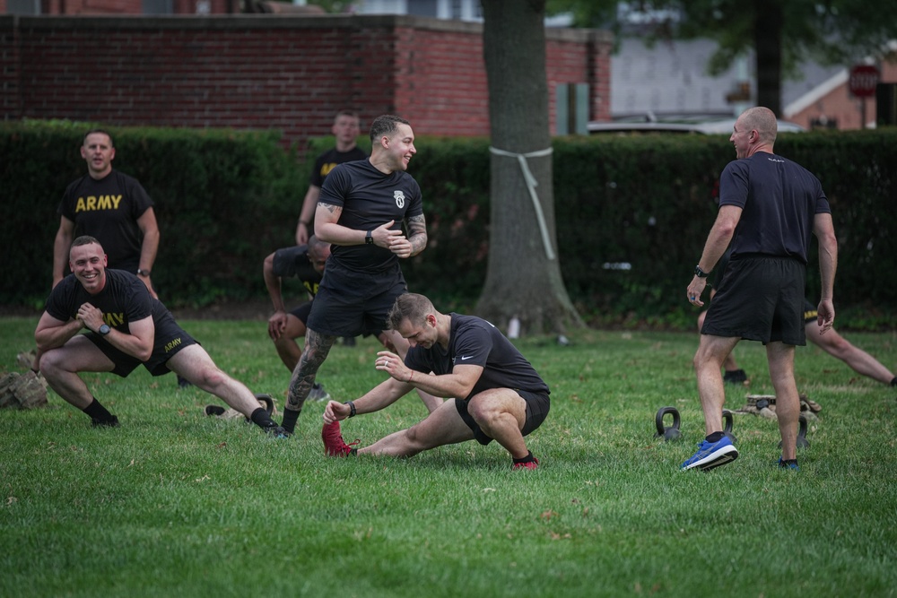 A group of U.S. Army soldiers and personnel are conducting an outdoor physical training session on a grass field. They are wearing black Army PT shirts and shorts, engaged in stretching and warm-up exercises. The setting appears to be a military installation or base, with brick buildings and hedgerows visible in the background.