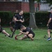 A group of U.S. Army soldiers and personnel are conducting an outdoor physical training session on a grass field. They are wearing black Army PT shirts and shorts, engaged in stretching and warm-up exercises. The setting appears to be a military installation or base, with brick buildings and hedgerows visible in the background.