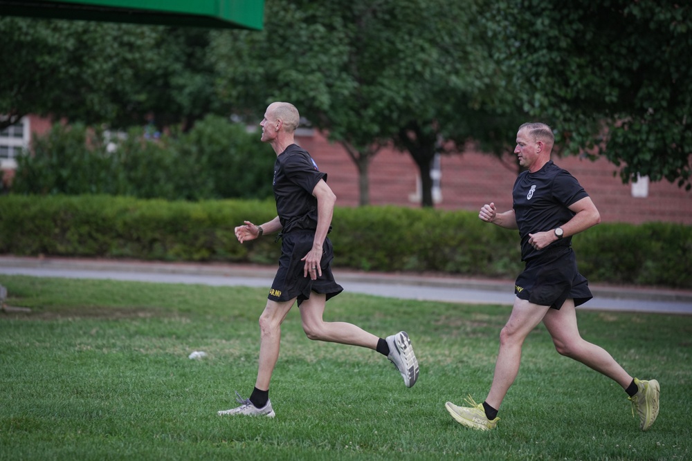 Two men in matching black Army PT (Physical Training) uniforms are running across a grass field. Both wear black shorts with yellow 'ARMY' lettering and black t-shirts with military insignia. The setting appears to be a military installation or base, with brick buildings and manicured hedges visible in the background.