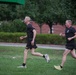 Two men in matching black Army PT (Physical Training) uniforms are running across a grass field. Both wear black shorts with yellow 'ARMY' lettering and black t-shirts with military insignia. The setting appears to be a military installation or base, with brick buildings and manicured hedges visible in the background.