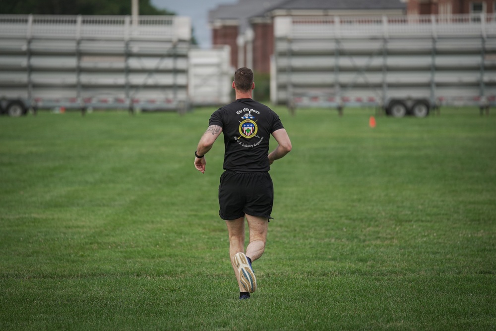 A man wearing a black 'The Old Guard - 3rd U.S. Infantry Regiment' t-shirt and black shorts is running away from the camera across a well-maintained grass field. The setting appears to be a military installation or sports complex, with bleachers/grandstands and trailers visible in the background. The image is taken from behind, capturing the subject mid-stride.