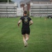 A man wearing a black 'The Old Guard - 3rd U.S. Infantry Regiment' t-shirt and black shorts is running away from the camera across a well-maintained grass field. The setting appears to be a military installation or sports complex, with bleachers/grandstands and trailers visible in the background. The image is taken from behind, capturing the subject mid-stride.