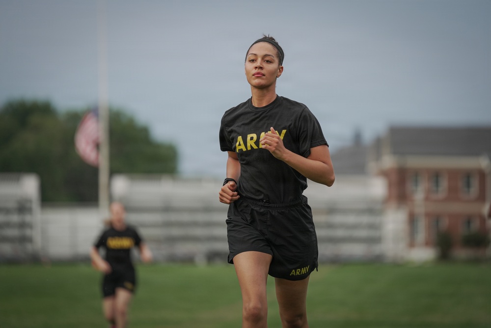 A female soldier in U.S. Army physical training (PT) uniform is running on a grassy field, likely during a fitness test or training exercise. She is wearing the standard black Army PT shirt and shorts with gold 'ARMY' lettering. A second soldier in identical PT uniform is visible in the blurred background, along with an American flag on a flagpole and a brick building.