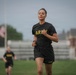 A female soldier in U.S. Army physical training (PT) uniform is running on a grassy field, likely during a fitness test or training exercise. She is wearing the standard black Army PT shirt and shorts with gold 'ARMY' lettering. A second soldier in identical PT uniform is visible in the blurred background, along with an American flag on a flagpole and a brick building.