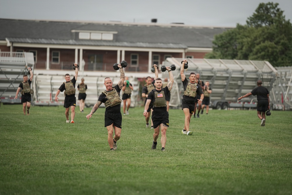 A group of military personnel in black PT uniforms and tactical plate carriers are conducting an outdoor physical training exercise on a grass field. The soldiers are running while holding kettlebells overhead with one arm, indicating a functional fitness or combat readiness drill. Bleachers and a brick building are visible in the background, suggesting a military installation or training facility.