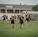 A group of military personnel in black PT uniforms and tactical plate carriers are conducting an outdoor physical training exercise on a grass field. The soldiers are running while holding kettlebells overhead with one arm, indicating a functional fitness or combat readiness drill. Bleachers and a brick building are visible in the background, suggesting a military installation or training facility.