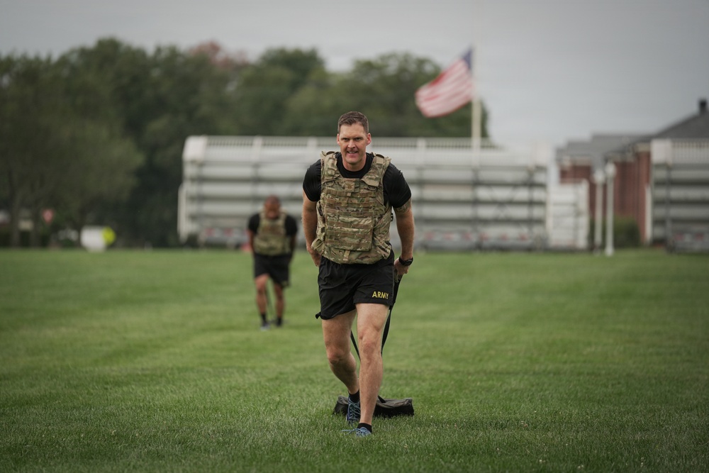 A U.S. Army soldier in the foreground is performing a physical training exercise on a grass field, wearing a tactical/plate carrier vest over a black t-shirt and Army-branded shorts. A second soldier in similar attire is visible in the background. An American flag flies on a flagpole behind them, with what appears to be a military installation building visible to the right.