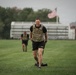 A U.S. Army soldier in the foreground is performing a physical training exercise on a grass field, wearing a tactical/plate carrier vest over a black t-shirt and Army-branded shorts. A second soldier in similar attire is visible in the background. An American flag flies on a flagpole behind them, with what appears to be a military installation building visible to the right.