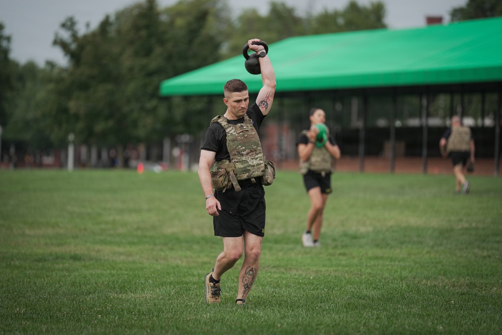 A man in military-style tactical gear performs a kettlebell overhead carry on a grass field, likely during a fitness competition or military physical training event. He is wearing a camouflage plate carrier vest, black shorts, and athletic shoes, with visible tattoos on his arm and leg. Several other participants are visible in the background performing similar exercises near a green canopy tent.