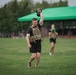 A man in military-style tactical gear performs a kettlebell overhead carry on a grass field, likely during a fitness competition or military physical training event. He is wearing a camouflage plate carrier vest, black shorts, and athletic shoes, with visible tattoos on his arm and leg. Several other participants are visible in the background performing similar exercises near a green canopy tent.