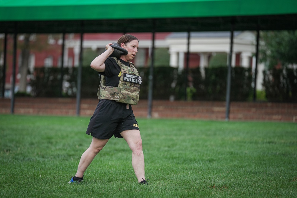A female soldier wearing a Military Police tactical vest over a camouflage uniform top and black Army shorts is performing a kettlebell exercise on a grass field. She appears to be executing a kettlebell clean or press movement, holding the weight at shoulder height. The setting appears to be a military installation based on the brick buildings and structured grounds visible in the background.