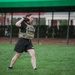 A female soldier wearing a Military Police tactical vest over a camouflage uniform top and black Army shorts is performing a kettlebell exercise on a grass field. She appears to be executing a kettlebell clean or press movement, holding the weight at shoulder height. The setting appears to be a military installation based on the brick buildings and structured grounds visible in the background.