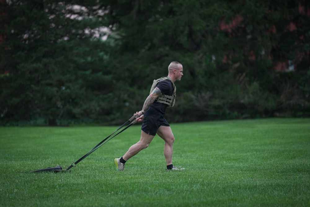 A muscular, short-haired male athlete is performing a sled drag exercise on a well-maintained grass field. He is wearing a tactical/weighted vest over a black t-shirt, black shorts, and athletic shoes, pulling a weighted sled behind him using resistance straps. The image is taken during overcast or early morning/evening lighting conditions with dense green trees in the background.