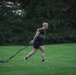 A muscular, short-haired male athlete is performing a sled drag exercise on a well-maintained grass field. He is wearing a tactical/weighted vest over a black t-shirt, black shorts, and athletic shoes, pulling a weighted sled behind him using resistance straps. The image is taken during overcast or early morning/evening lighting conditions with dense green trees in the background.