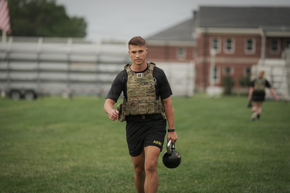 A male soldier in U.S. Army physical training attire walks across a grass field carrying a kettlebell in his right hand. He is wearing a multicam plate carrier vest over a black t-shirt and black Army-branded shorts. A second person in similar attire is visible in the background near a brick building.