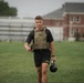 A male soldier in U.S. Army physical training attire walks across a grass field carrying a kettlebell in his right hand. He is wearing a multicam plate carrier vest over a black t-shirt and black Army-branded shorts. A second person in similar attire is visible in the background near a brick building.