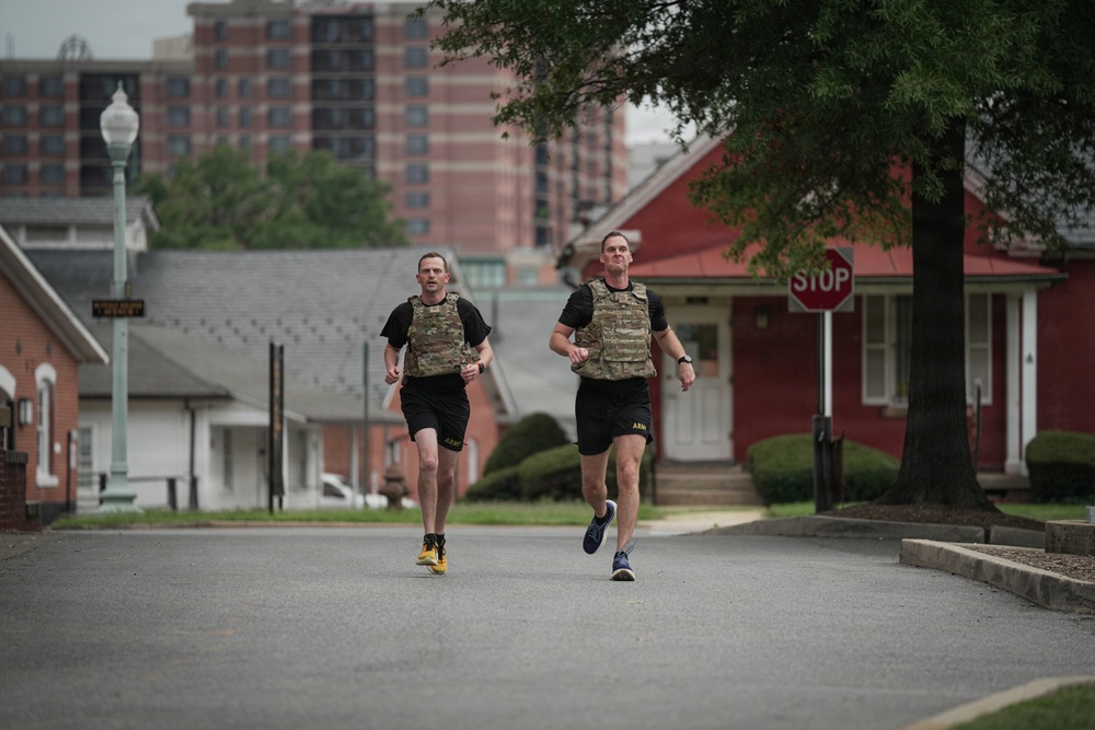 Two individuals wearing U.S. Army physical training uniforms with camouflage plate carriers/weighted vests are running down a paved road in what appears to be a military installation or base. Both men are wearing black shorts with 'ARMY' lettering and running shoes, suggesting this is an official physical fitness training exercise. The background shows low brick buildings, a stop sign, a street lamp, and a taller brick building in the distance.