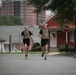 Two individuals wearing U.S. Army physical training uniforms with camouflage plate carriers/weighted vests are running down a paved road in what appears to be a military installation or base. Both men are wearing black shorts with 'ARMY' lettering and running shoes, suggesting this is an official physical fitness training exercise. The background shows low brick buildings, a stop sign, a street lamp, and a taller brick building in the distance.