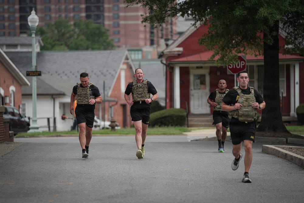 Four individuals wearing military-style tactical plate carriers and black athletic uniforms are running down a paved road in what appears to be a military installation or base housing area. The runners are wearing matching black shorts and shirts with what appears to be 'Army' branding, suggesting this is a military physical training exercise. The setting includes red brick residential buildings, a stop sign, a street lamp, and mature trees in the background.