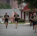 Four individuals wearing military-style tactical plate carriers and black athletic uniforms are running down a paved road in what appears to be a military installation or base housing area. The runners are wearing matching black shorts and shirts with what appears to be 'Army' branding, suggesting this is a military physical training exercise. The setting includes red brick residential buildings, a stop sign, a street lamp, and mature trees in the background.
