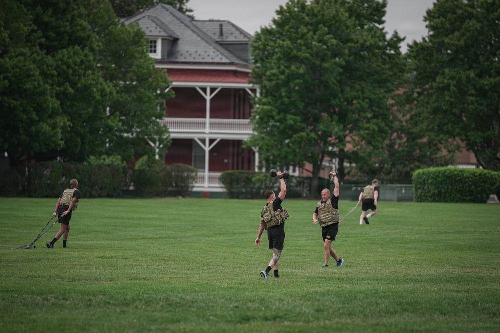 A group of individuals in black athletic wear and tactical/plate carrier vests are performing outdoor fitness training on a large grass field. Two central figures are performing overhead kettlebell carries while walking, and at least two others in the background appear to be dragging weighted sleds. The setting appears to be a military installation or institutional campus, evidenced by the historic red-brick building with white porches visible in the background.