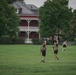 A group of individuals in black athletic wear and tactical/plate carrier vests are performing outdoor fitness training on a large grass field. Two central figures are performing overhead kettlebell carries while walking, and at least two others in the background appear to be dragging weighted sleds. The setting appears to be a military installation or institutional campus, evidenced by the historic red-brick building with white porches visible in the background.