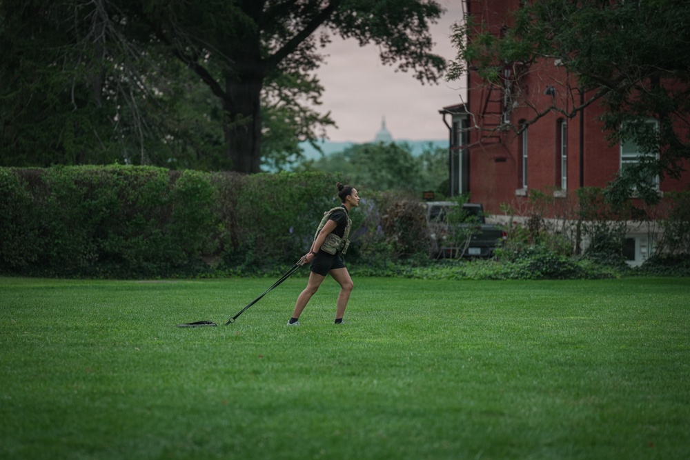 A woman is performing an outdoor fitness workout on a large green lawn, pulling a weighted sled using resistance straps. She is wearing a tactical/weighted vest over black athletic clothing. The setting appears to be near a red brick institutional building, likely during early morning or evening hours based on the muted sky.
