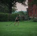 A woman is performing an outdoor fitness workout on a large green lawn, pulling a weighted sled using resistance straps. She is wearing a tactical/weighted vest over black athletic clothing. The setting appears to be near a red brick institutional building, likely during early morning or evening hours based on the muted sky.